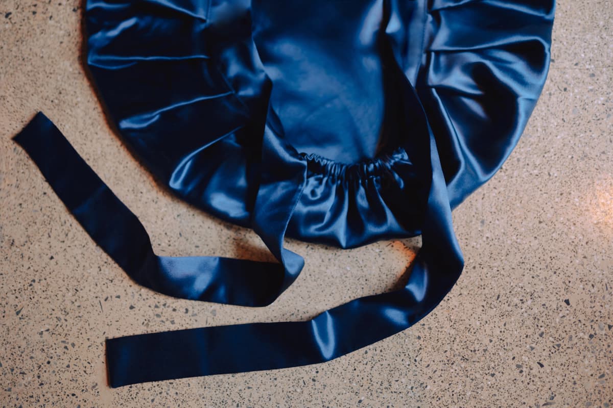 Dark navy silk All Rounder bonnet laid flat on a terrazzo floor, showing its gathered elasticated edge and long adjustable silk ties.
