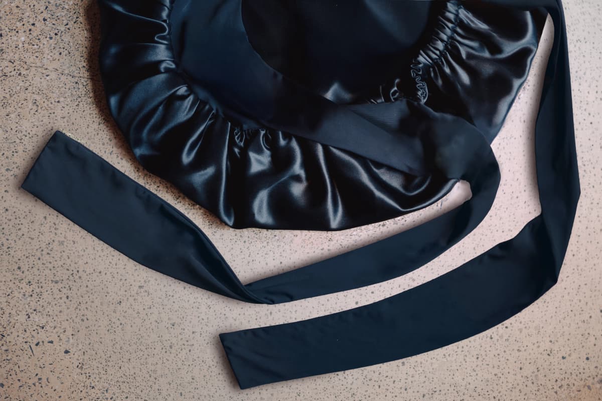 Dark navy silk Secure Wrap bonnet laid flat on a terrazzo floor, showing its gathered elasticated edge and long adjustable wrap ties.