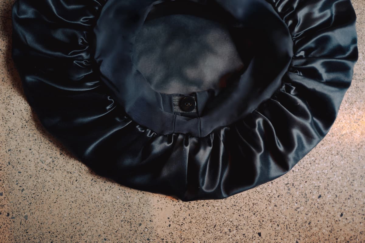 Black silk Smooth Fit bonnet laid flat on a terrazzo floor, showing its gathered elasticated edge and button-adjustable band.