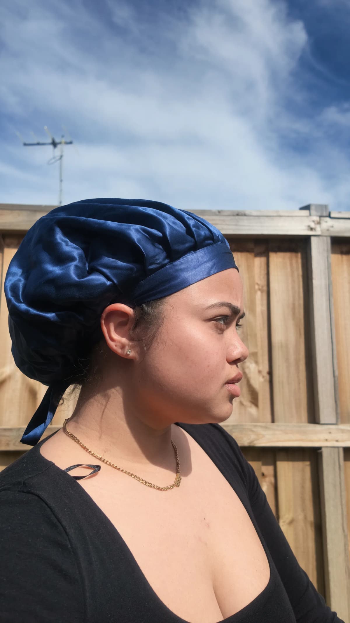 Side profile of a customer wearing the navy Kova Silk All Rounder bonnet outdoors in front of a wooden fence.