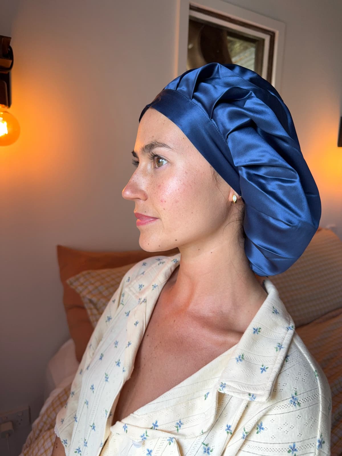 Side profile of a person wearing a navy silk bonnet and a floral pyjama top, indoors by warm lamplight.