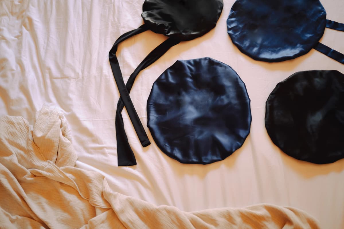 The four Kova Silk bonnets laid out on soft pink bedding — one black and three navy silk bonnets with ties, styled with a textured throw.