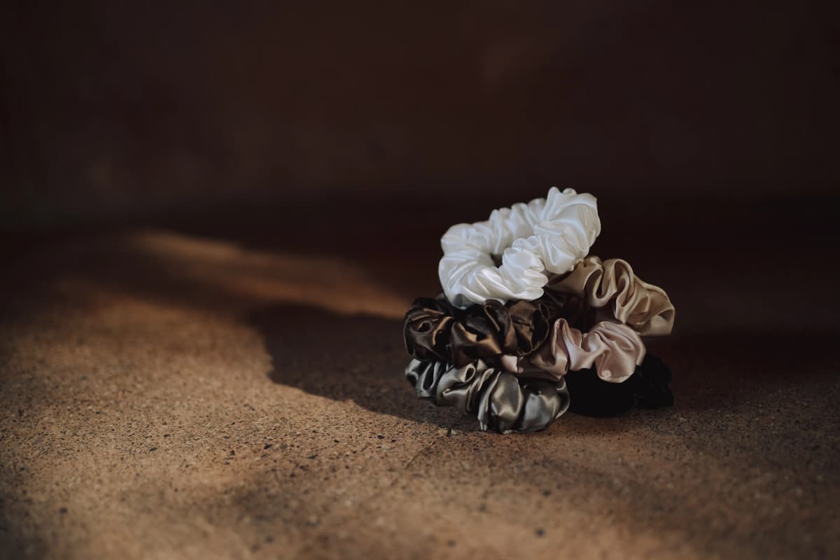 A cluster of Kova Silk scrunchies in ivory, taupe, chocolate, pewter and black resting in warm natural light