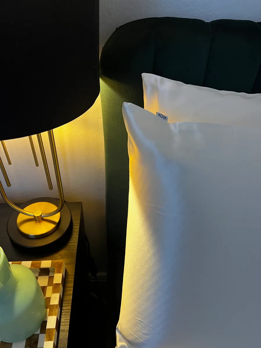 A white Kova Silk pillowcase on a bed beside a brass bedside lamp in warm light